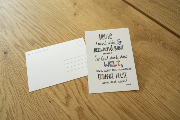weiße Postkarte mit bunter Schrift "Heute tanzt dein Tag hellwach & bunt drapiert sol laut durch deine Welt, dass selbst der trennendste Gedanke heute seinen Mut verliert!"