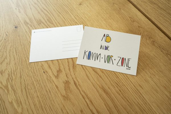 weiße Postkarte mit bunter Schrift "Ab in die Komm vor Zone"