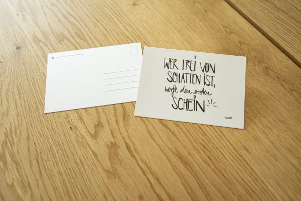 weiße Postkarte mit bunter Schrift "Wer frei von Schatten ist, werde den ersten Schein"